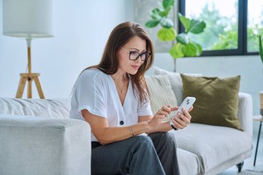 Orta yaşlı, rahat, gülümseyen mutlu bir kadın evde koltukta uzanmış akıllı telefon mesajları okuyor. Boş zaman iletişimi için mobil uygulamalar iş eğitimi teknoloji yaşam tarzı insanlar kavramı