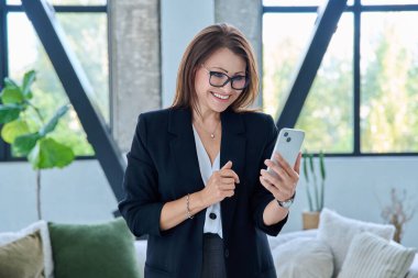 Ofiste koltuklu akıllı telefon kullanan orta yaşlı bir iş kadını. İş, iş, teknoloji, iletişim, mobil dijital uygulamalar kavramı