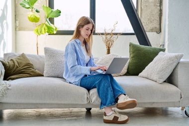 Genç bir kadının portresi kanepede oturuyor ve dizüstü bilgisayarda ev içinde daktilo yazıyor. Üniversite öğrencisi, internetten sosyal medya blogu alışverişini rahatlatmak için sınava hazırlanıyor.