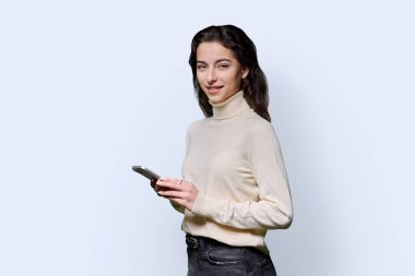 Beyaz stüdyo arka planında akıllı telefonu tutan gülümseyen genç kızın portresi. Genç bayan cep telefonu kullanıyor, kameraya bakıyor. Teknoloji, eğitim, eğlence ve iletişim için mobil uygulamalar