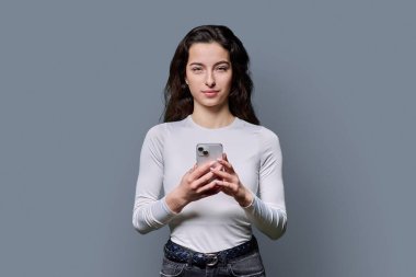 Gri arka planda akıllı telefonu tutan gülümseyen genç kızın portresi. Genç bayan cep telefonu kullanıyor, kameraya bakıyor. Teknoloji, mobil eğitim uygulamaları, boş zaman, iletişim