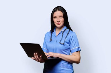 Diz üstü bilgisayarı tutan gülümseyen kadın hemşire, beyaz stüdyo arka planındaki kameraya bakıyor. Mobil uygulamalar tıbbi hizmetler, sağlık, profesyonel yardım, tıbbi bakım konsepti teknolojileri