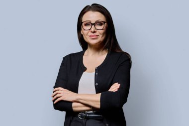 Gri arka planda çapraz kollu, kendine güvenen olgun bir kadının portresi. Gözlüklü, siyah elbiseli güzel bir kadın stüdyoda poz veriyor. Güzellik, sağlık, olgunluk, insan kavramı