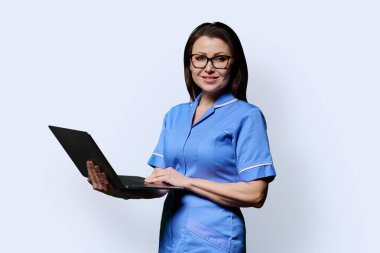 Diz üstü bilgisayarı tutan gülümseyen kadın hemşire, beyaz stüdyo arka planındaki kameraya bakıyor. Mobil uygulamalar tıbbi hizmetler, sağlık, profesyonel yardım, tıbbi bakım konsepti teknolojileri