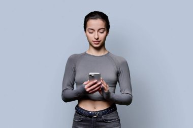 Gri arka planda akıllı telefonu tutan gülümseyen genç kızın portresi. Genç bir bayan cep telefonu kullanıyor, telefona bakıyor. Teknoloji, mobil eğitim uygulamaları, boş zaman, iletişim