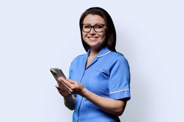 Beyaz stüdyo arka planında akıllı telefonlu gülümseyen kadın hemşire portresi. Mobil uygulamalar, tıbbi hizmetlerdeki teknolojiler, sağlık, profesyonel yardım, tıbbi bakım kavramı