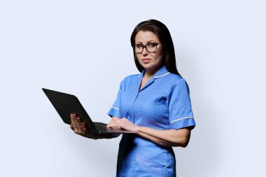 Ciddi bir bayan hemşire dizüstü bilgisayar tutuyor, beyaz stüdyo arka planındaki kameraya bakıyor. Mobil uygulamalar tıbbi hizmetler, sağlık, profesyonel yardım, tıbbi bakım konsepti teknolojileri
