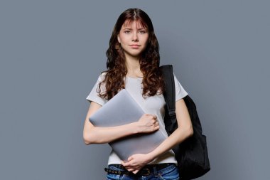 Sırt çantalı, dizüstü bilgisayarı gri arka planda kameraya bakan güzel, gülümseyen genç bir kızın portresi. Eğitim, okul, öğrenme, teknoloji, okul çocukları kavramı