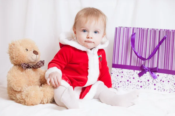 Um bebê-menina vestida vermelho fantasia vestido de Papai Noel com saco de papel violeta para presentes e grande ursinho de pelúcia — Fotografia de Stock