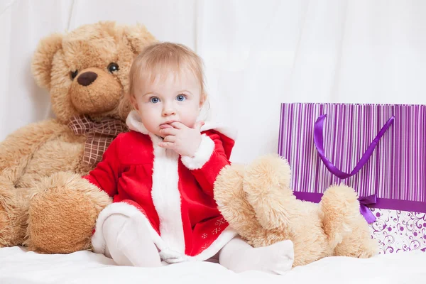 Una pequeña niña vestida de rojo elegante vestido de Santa Claus con bolsa de papel violeta para regalos y oso de peluche grande — Foto de Stock