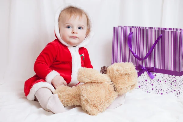En liten flicka klädd röd fancy jultomten klänning med violett papperspåse för presenter och stora nallebjörn — Stockfoto