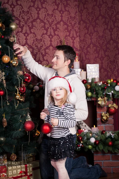 Far och dotter belysa en julgran av stora ljusa röda och gyllene bollar. En flicka med Santa Claus röd hatt på huvudet håller en röd boll. Det finns en öppen spis bakom dem.. — Stockfoto