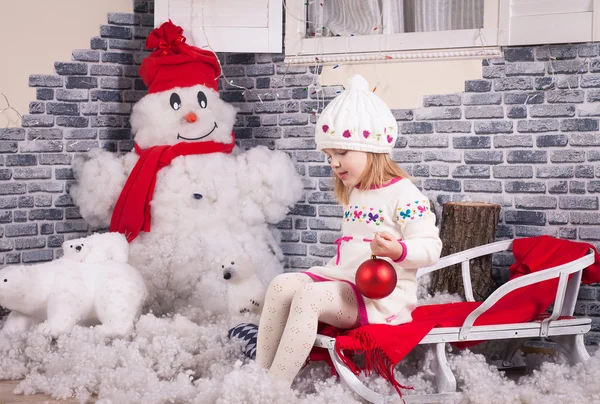 Une petite fille sur le traîneau avec une boule de sapin rouge dans les mains. Dos décoré par imitation de neige et bonhomme de neige près du mur de briques — Photo