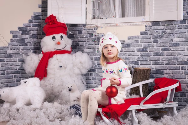 Une petite fille sur le traîneau avec une boule de sapin rouge dans les mains. Dos décoré par imitation de neige et bonhomme de neige près du mur de briques — Photo