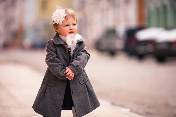 Ett porträtt av söt liten baby flicka i grå kappa på gatan i gamla staden med rolig leende — Stockfoto