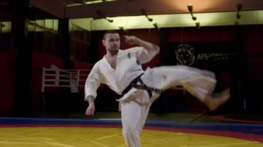 Karate ustası spor salonunda tatamiye zıplayarak bacak tekmeliyor.