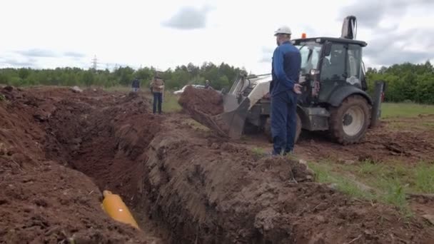 Employees stand near excavator and trench with yellow pipe — Stock Video