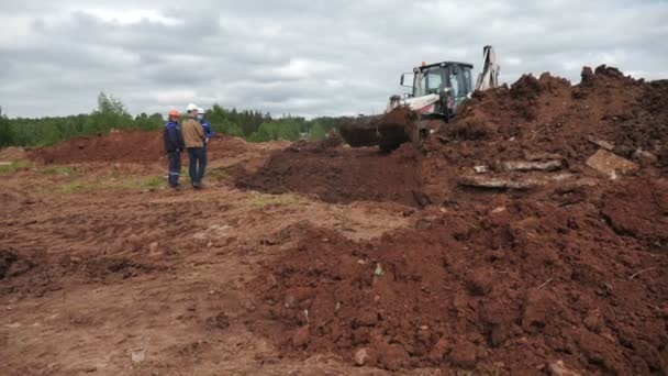 La excavadora lleva la suciedad a la zanja bajo control de los constructores — Vídeo de stock