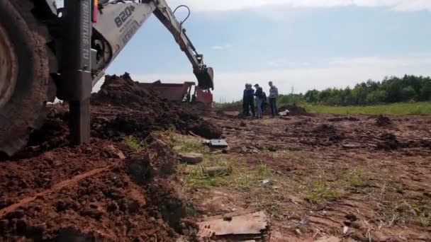 Экскаватор копает яму в грязи, пока рабочие контролируют процесс — стоковое видео
