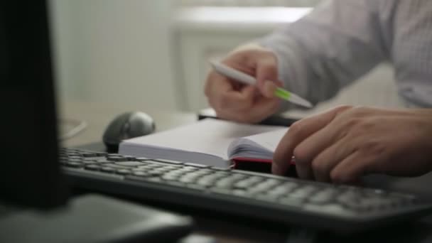 Joven escribe en cuaderno sentado cerca de la computadora en la oficina — Vídeos de Stock