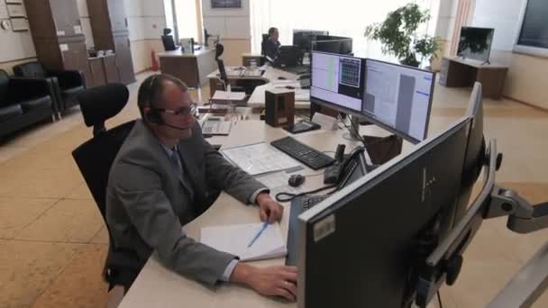 Человек с гарнитурой сидит за столом с мониторами в комнате управления — стоковое видео