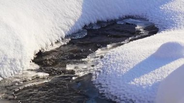 Dar akıntı beyaz vadiyi keser ve canlı bir hareket çizgisi yaratır. Soğuk eriyen su, yumuşak kanalı karla oyar ve karanlık toprağı ortaya çıkarır.
