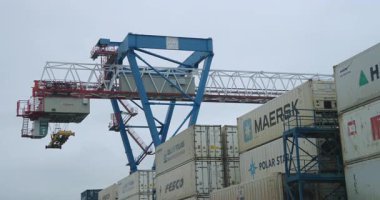 ALMATY, KAZAKHSTAN - 09 Ekim 2025: Soluk bulutlu gökyüzü altında parlayan işaretlenmiş kutuların üzerinde yükselen konteyner vinci. Devasa çelik kirişler sakin deniz manzarasına hükmediyor