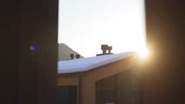 Kışın güneş doğarken perdelerin arasından karlı çatısı olan bir bina. Soğuk bir sabahta huzurlu bir dağ evinde uyanmak.