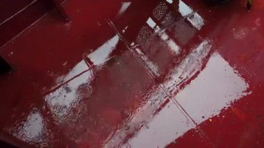 Metalik güverte yağmur damlaları mükemmel daireler oluştururken çiseleme altında parlar. Damlalar aynalı su birikintileriyle birleşir hipnotik deniz ritmi yaratır.