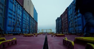 Parlak kasklı erkek işçi, yüksek konteynırlar arasında metal güvertede yürüyor. Sakin adımların ritmi sessiz deniz gücünü yansıtır.