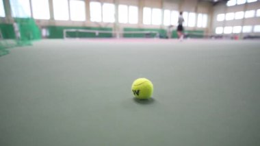 Tenis topu kapalı alandaki saha yüzeyine bağlıyken, bulanık sporcu uzun pencerelerin yanında ısınıyor. Sportif atmosfer odaklanma hazırlığını vurguluyor