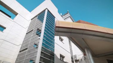 Modern cam kule, mavi gökyüzünün altındaki metal kaplı binanın yanında yükseliyor. Cantilevered tepe örtüsü, dikey pencere ızgarasını çerçeveleyen kanatlara bağlanır