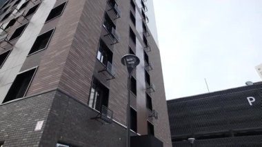 Ön planda yükselen ince sokak lambaları, arkadaki büyük yerleşim hacmini vurguluyor. Kompozisyon dikey hakimiyeti vurgular