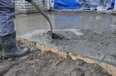 Kauçuk çizmeli bir inşaatçının, tahta bir levha için ıslak betonu ahşap bir kalıba dökerek ve dökerek yakın plan çekimi. İnşaat alanı somutlaştırma ve temel çalışmaları
