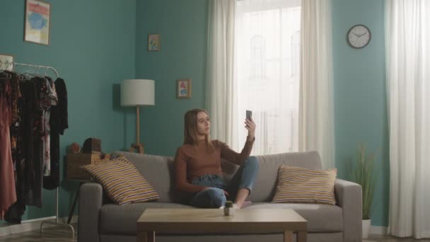 Молодая девушка сидит на диване и разговаривает по телефону по видеосвязи — стоковое видео