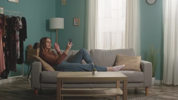 Genç kız kanepede uzanıyor ve video bağlantısıyla telefonla konuşuyor. — Stok video
