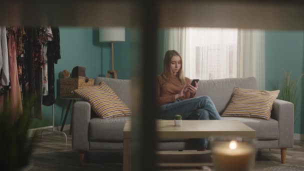 Jeune fille assise sur le canapé et téléphone parlant via lien vidéo — Video