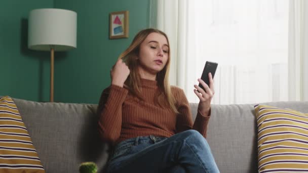 Молодая девушка сидит на диване и разговаривает по видеосвязи — стоковое видео
