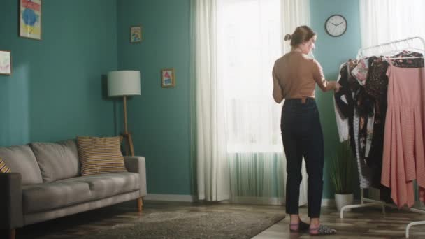 Jonge vrouw neemt kleren aan een hanger en neemt ze uit de kamer — Stockvideo
