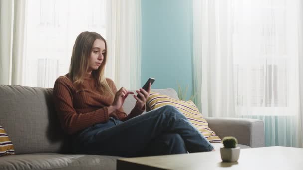 Junges Mädchen sitzt auf dem Sofa und hält ein Handy in der Hand — Stockvideo