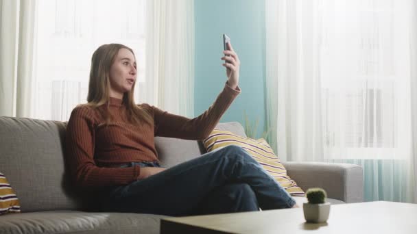 Chica joven está sentada en un sofá y hablando en una videollamada — Vídeos de Stock