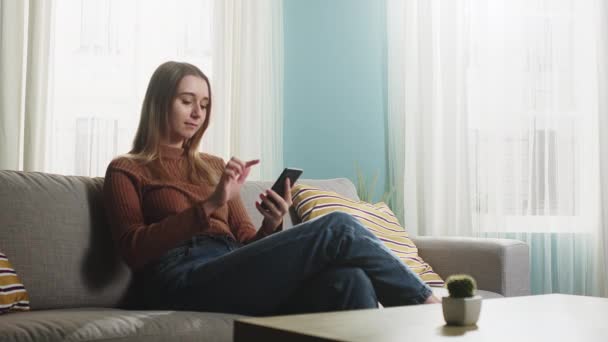 Junges Mädchen sitzt auf dem Sofa und hält ein Handy in der Hand — Stockvideo