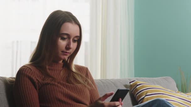 Молода дівчина сидить на дивані з телефоном в руках — стокове відео