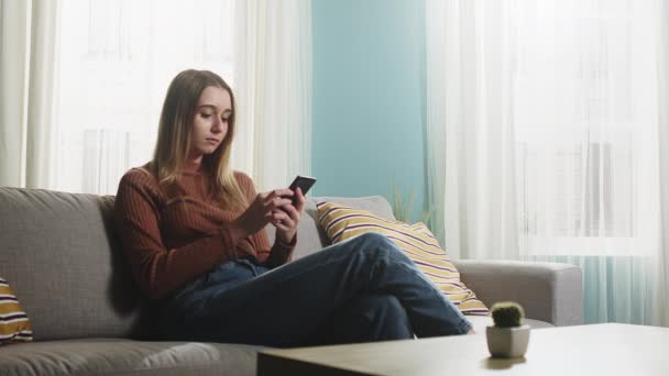 Молодая девушка сидит на диване с телефоном в руках — стоковое видео