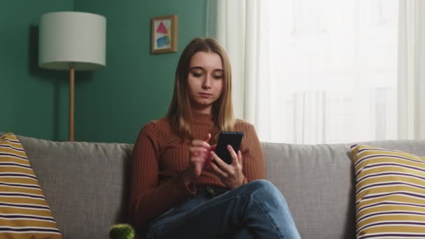 Jovem sentada no sofá com um telefone em suas mãos — Vídeo de Stock