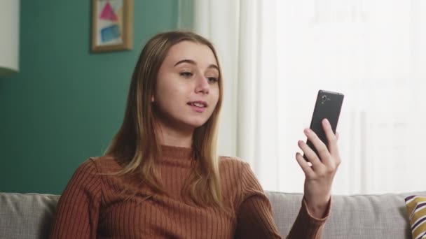 Молода дівчина сидить на дивані і розмовляє на відеодзвінок — стокове відео