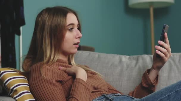 Una joven está tumbada en el sofá y hablando en una videollamada — Vídeos de Stock