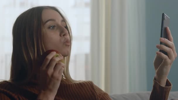 Schöne Mädchen mit rotem Apfel sitzt auf Sofa und macht Selfie auf Gadget — Stockvideo