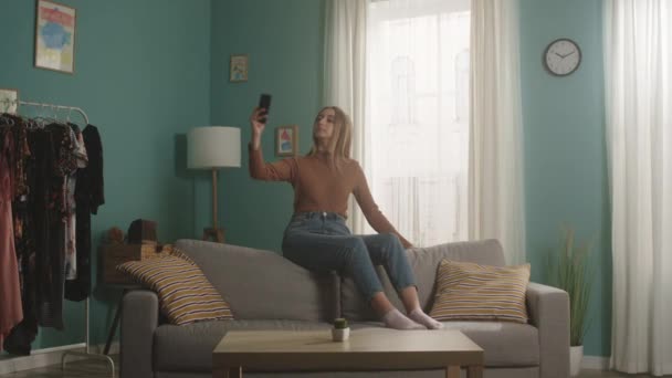 Девочка садится на спинку дивана и делает селфи — стоковое видео