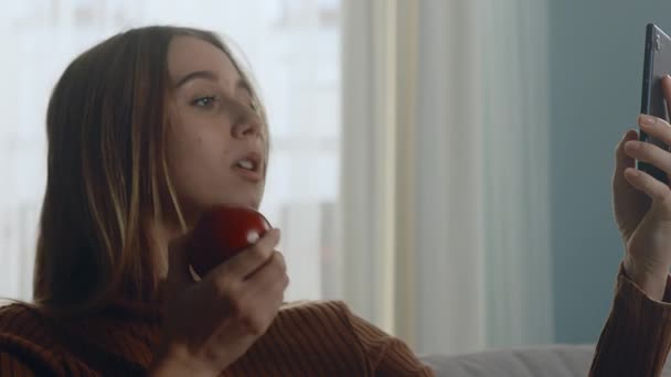 Mooie vrouw met rode appel zit op de bank en maakt selfie op gadget — Stockvideo